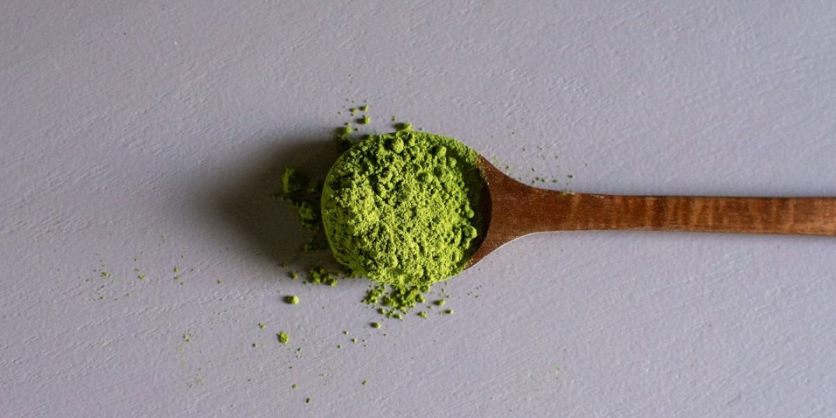 Cucharada de madera llena de polvo verde de té matcha sobre superficie blanca con algunos granos dispersos