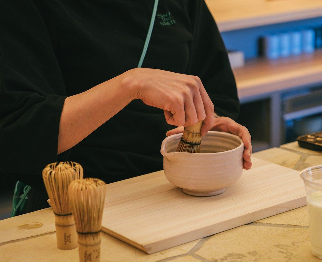 Persona batiendo matcha en polvo con un batidor de bambú dentro de un cuenco de cerámica blanca sobre una tabla de madera