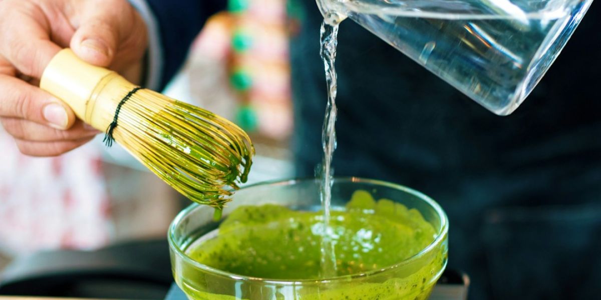 Persona vertiendo agua en un cuenco con matcha verde en polvo mezclado con un batidor de bambú amarillo