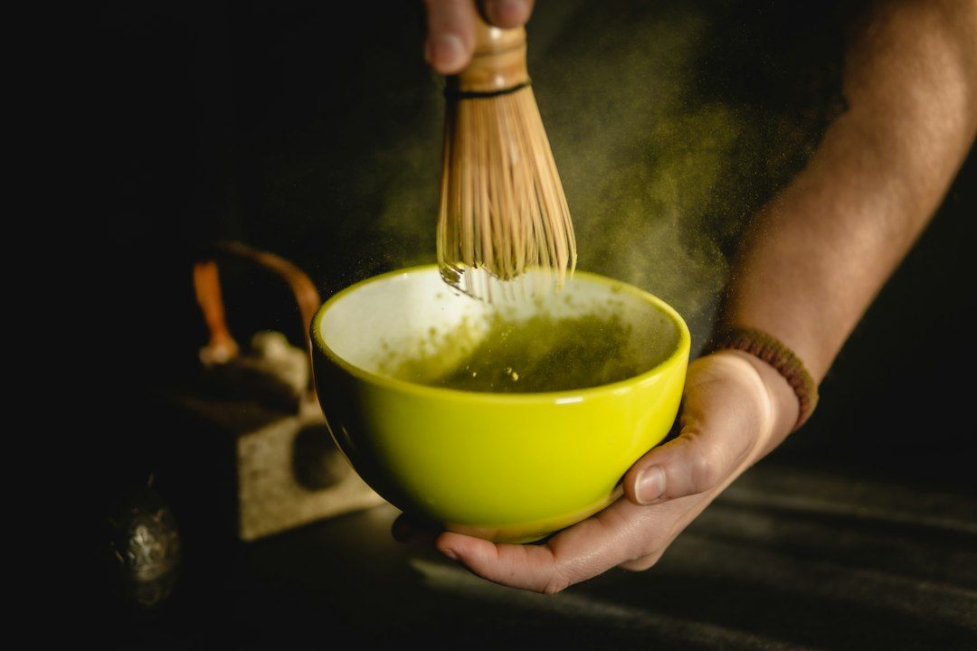 Manos preparando matcha con batidor tradicional de bambú en cuenco amarillo sobre fondo oscuro