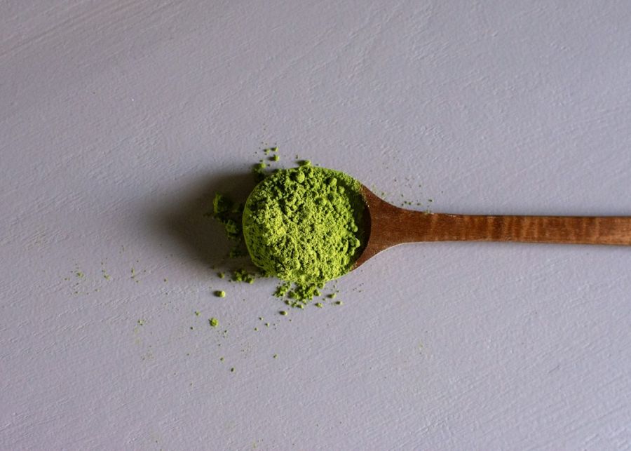 Cucharada de madera llena de polvo verde de té matcha sobre superficie blanca con algunos granos dispersos