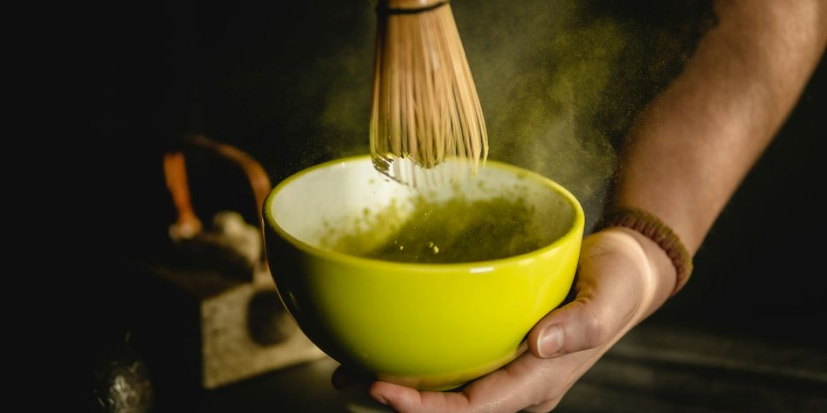 Manos preparando matcha con batidor tradicional de bambú en cuenco amarillo sobre fondo oscuro