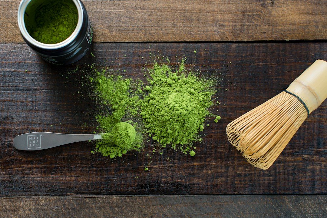 Polvo de matcha verde sobre tabla de madera con cucharilla de medición y batidor de bambú para preparar matcha en ayunas