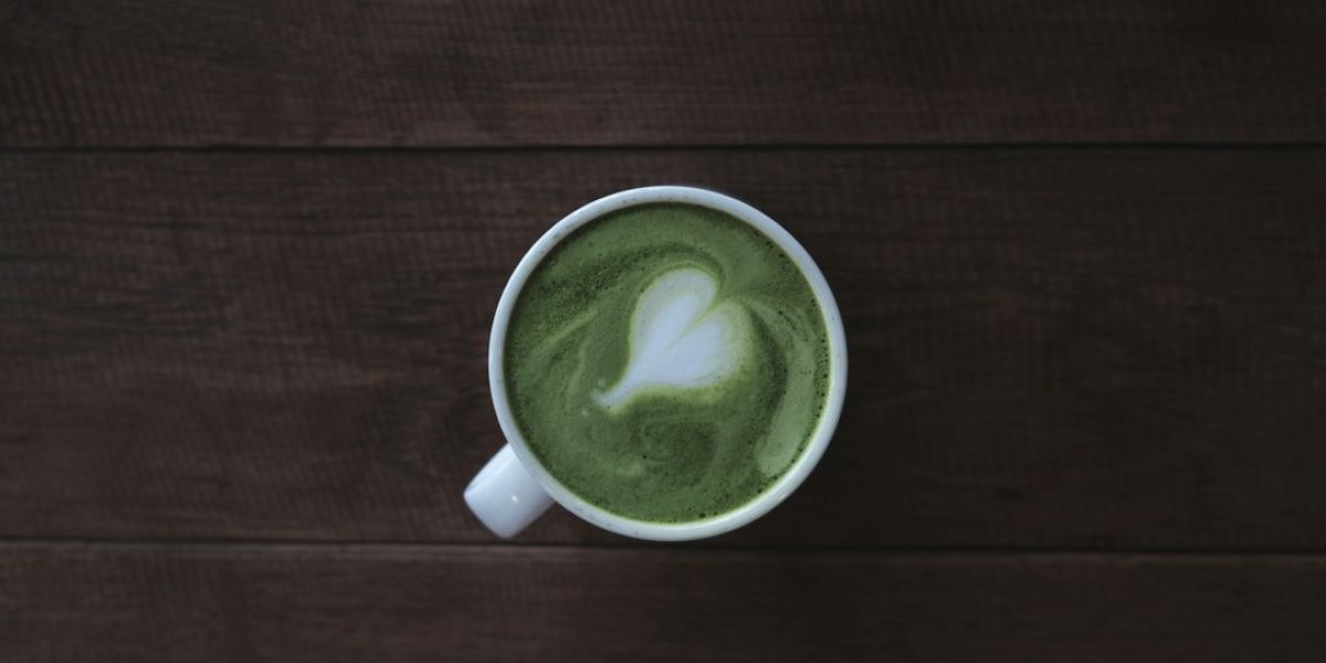 Taza blanca con matcha latte de color verde intenso con latte art en forma de hoja sobre superficie de madera oscura