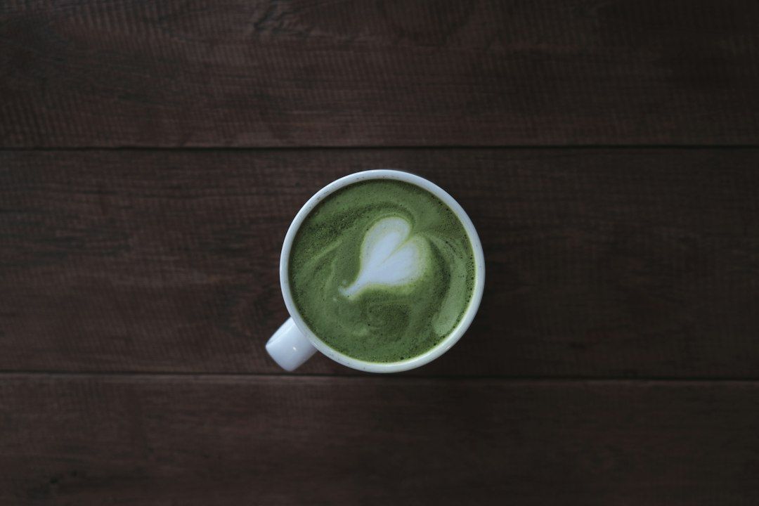 Taza blanca con matcha latte de color verde intenso con latte art en forma de hoja sobre superficie de madera oscura