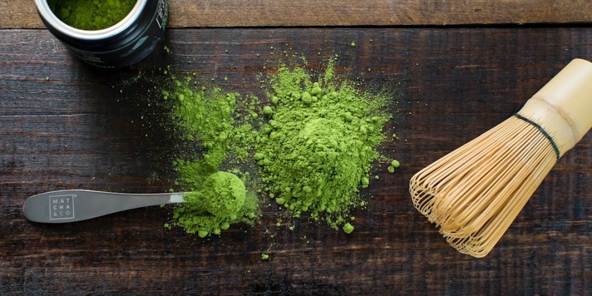 Polvo de matcha verde sobre tabla de madera con cucharilla de medición y batidor de bambú para preparar matcha en ayunas