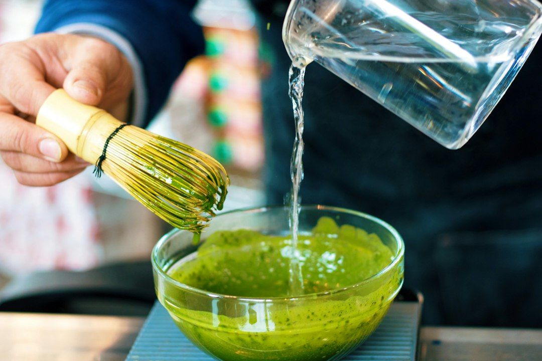 Persona vertiendo agua en un cuenco con matcha verde en polvo mezclado con un batidor de bambú amarillo
