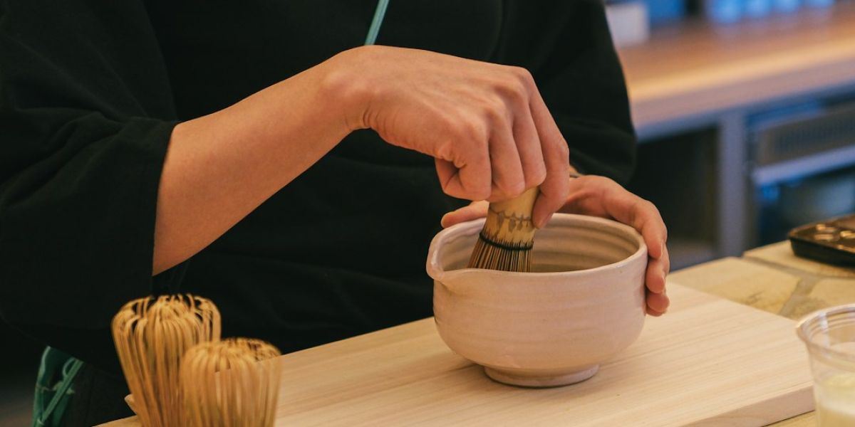 Persona batiendo matcha en polvo con un batidor de bambú dentro de un cuenco de cerámica blanca sobre una tabla de madera