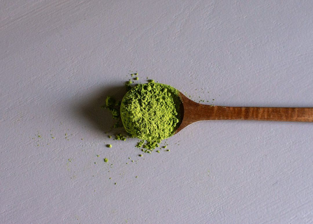 Cucharada de madera llena de polvo verde de té matcha sobre superficie blanca con algunos granos dispersos